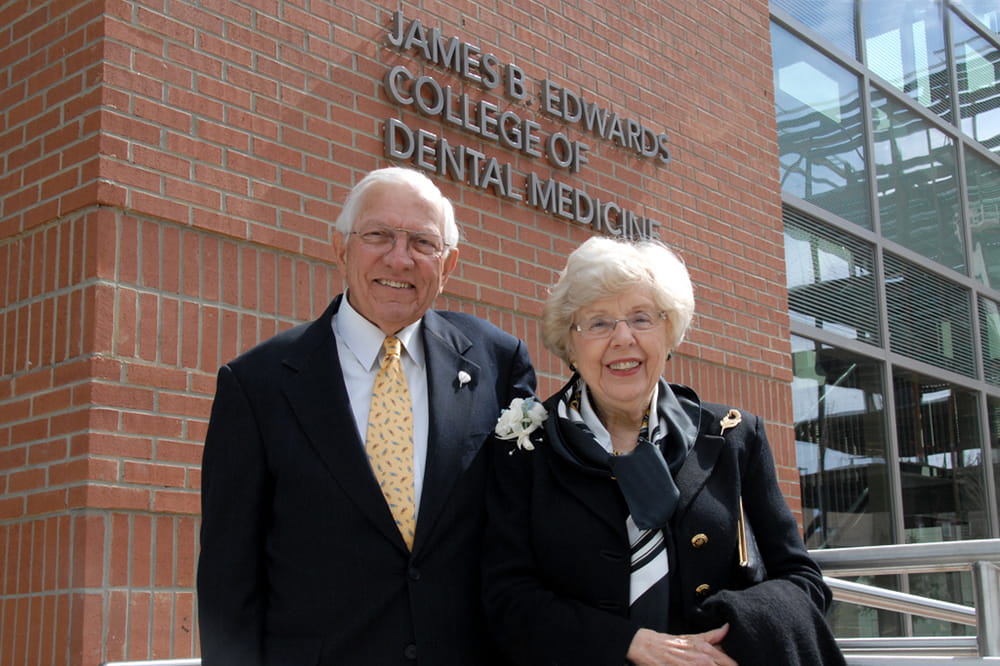 A man wearing a suit and tie and a woman who is also in dress clothes stand in front of a brick building bearing the words James B. Edwards College of Dental Medicine.