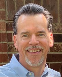 Man with gray hair and a goatee is wearing a blue shirt and smiling.